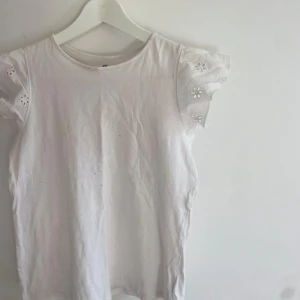 Vit blus med broderade ärmar H&M - Söt vit blus från H&M med korta volangärmar som har broderade blommor. Toppen har rund halsringning och är gjord i mjuk bomull. Små fläckar men inte jättesynliga!