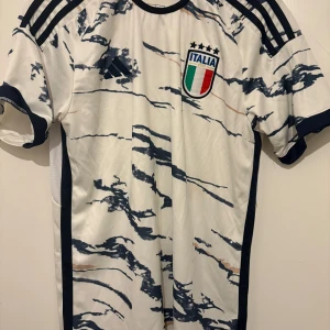 Italien fotbollströja Adidas S - Italiens landslagströja från Adidas i slim fit, vit med marinblå och beige mönsterdetaljer. Korta ärmar, broderat landslagsmärke och klassiska Adidas-ränder på axlarna. Tillverkad i lätt och ventilerande material.
