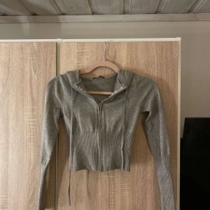 Grå croppad hoodie från Catania - Snygg grå croppad hoodie från Catania med dragkedja framtill och långa ärmar. Tröjan har ribbade detaljer vid midjan och ärmsluten samt en huva med snörning. Perfekt för en chill och trendig look.