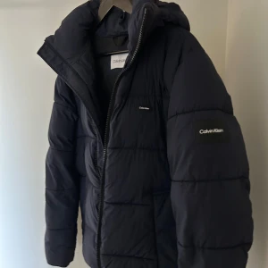 Mörkblå pufferjacka från Calvin Klein - Säljer en mörkblå pufferjacka från Calvin Klein med luva och diskret logga på bröst och arm. Jackan är använd en vinter och säljer den då den tyvärr blivit för liten. Nypris 2399