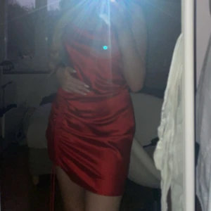 Röd satin klänning med snörning - Snygg röd kort klänning i satin med tunna axelband och draperad snörning längs ena sidan. Klänningen har en lätt glansig finish och en smickrande passform som framhäver figuren. Perfekt för dig som vill sticka ut med en trendig look.Denna säljs inte längre!