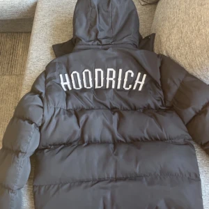 Svart pufferjacka från Hoodrich - Svart pufferjacka från Hoodrich med stor vit logga på ryggen och mindre logga på bröstet. Jackan har huva, dragkedja framtill och två sidofickor. Materialet är syntet och jackan är vadderad för extra värme. Perfekt för kalla dagar.