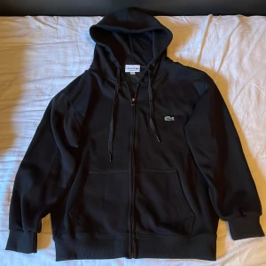 Svart hoodie från Lacoste med dragkedja - Svart hoodie från Lacoste i regular fit med klassisk krokodillogga på bröstet. Tröjan har huva med snörning, hel dragkedja framtill och två fickor. Tillverkad i mjuk bomullsmix, perfekt för chill dagar. Långa ärmar och ribbade muddar för extra komfort.                          Helt ny, har bara testat den 1 gång. Säljer iväg den för att den var för liten för mig.
