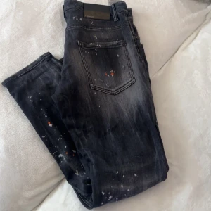 Dsq2 jeans  - Säljer ett par svarta jeans från Dsquared2 med coola slitningar och färgstänk i vitt och orange. Jeansen har en smal passform, klassisk femficksdesign och detaljer som ger en edgy streetkänsla. Perfekta för dig som vill sticka ut med din stil. Sitter som 30-32 ( 48 i storlek dsq2)