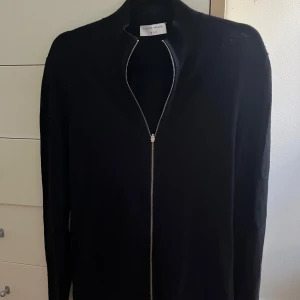 Svart kofta med dragkedja Tiger of Sweden - Svart, stilren kofta från Tiger of Sweden i storlek S. Koftan har hel dragkedja framtill, ribbad krage och muddar samt diskret broderad 1903 vid nederkanten. Mycket bra skick.