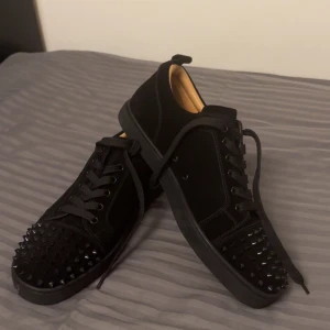 Christian Louboutin svarta sneakers med nitar - Säljer ett par svarta sneakers från Christian Louboutin i mocka med coola svarta nitar på tån och den ikoniska röda sulan. Skorna har snörning och en rund tå, perfekt för dig som vill sticka ut med en lyxig och edgy stil.