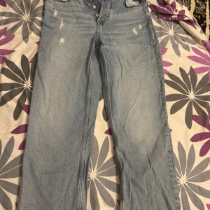 Ljusblå raka jeans från Zara - Ljusblå jeans från Zara med raka ben och hög midja. Jeansen har slitna detaljer framtill, klassiska fem fickor och knäppning med silverfärgade knappar. Tillverkade i bomullsdenim för en avslappnad vibe.