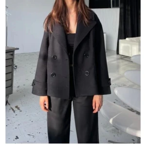 Svart kappa med knappar - Stilren svart kappa med bred krage och dubbelknäppning framtill. Jackan har vida ärmar och stora knappar både fram och vid ärmslut. Perfekt för dig som gillar en klassisk och cool look.