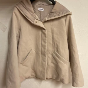 Beige kappa med huva från Bubbleroom - Stilren beige kappa från Bubbleroom med huva och guldig dragkedja. Jackan har två snedställda fickor framtill och är fodrad på insidan. Materialet är mjukt och ulligt, perfekt för kyliga dagar. Enkel och clean design som passar till det mesta.