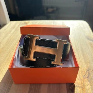 Svart bälte med guld spänne - Säljer ett svart bälte. Bältet är gjort i exklusivt skinn med vit kontrastsöm och levereras i original orange box. Perfekt accessoar för att lyfta vilken outfit som helst.