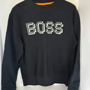 Svart sweatshirt från BOSS med pixeltryck - Svart sweatshirt från BOSS med stort pixelinspirerat BOSS-tryck på bröstet. Tröjan har rund hals, långa ärmar och ribbade muddar. Perfekt för dig som gillar streetwear och vill sticka ut med en cool grafisk detalj.