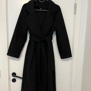 Svart lång kappa med bälte - Stilren svart kappa med klassisk krage och knytskärp i midjan. Lång modell med raka ärmar och snyggt fall. Perfekt för dig som vill ha en tidlös och elegant look.