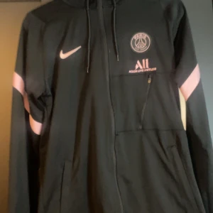 Svart PSG vindjacka från Nike S - Svart hoodie från Nike med Paris Saint-Germain-logga och rosa detaljer på ärmarna. Jackan har dragkedja, huva med snörning och Dri-FIT-material som andas. Perfekt för fotboll eller streetwear. Snyggt broderad PSG-emblem och Accor Live Limitless-tryck på bröstet.