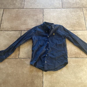 Mörkblå skjorta från Polo Ralph Lauren - Mörkblå skjorta från Polo Ralph Lauren i slim fit med klassisk krage och knappar framtill. Skjortan har lång ärm och ikonisk gul broderad logga på bröstet. Tillverkad i mjuk bomull och passar perfekt till en stilren look.