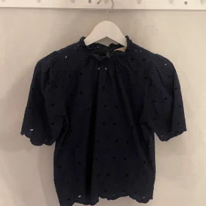 Mörkblå blus Holly & Whyte - Supersöt mörkblå blus från Holly & Whyte. Blusen har korta puffärmar, liten volang vid halsen och knäppning bak i nacken. 💞