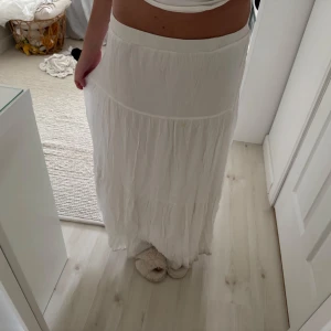 Vit långkjol  - Säljer en flowy vit långkjol med bred resår i midjan och volangdetaljer. Kjolen är tillverkad i tunt, lätt bomullstyg som ger en luftig känsla. Perfekt för varma dagar och enkel att styla med crop tops eller linnen. Har fläckar längst ned