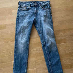 Blå jeans från Tommy Hilfiger - Snygga blå jeans från Tommy Hilfiger med klassisk femficksdesign och diskret logga på bakfickan. Jeansen har en rak passform och är tillverkade i slitstarkt bomullsjeans. Färgen är mellanblå med lätt slitningar för en cool, avslappnad look.