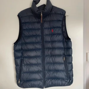 Blå dunväst från Polo Ralph Lauren - Snygg marinblå dunväst från Polo Ralph Lauren med hög krage, dragkedja framtill och två fickor med dragkedja. Klassisk quiltad design och röd broderad logga på bröstet. Perfekt för lager-på-lager och stilren look. Priset kan diskuteras! Mvh
