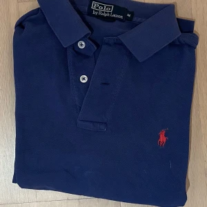 Marinblå pikétröja från Ralph Lauren - Klassisk marinblå pikétröja från Polo Ralph Lauren med röd broderad logga på bröstet. Tröjan har korta ärmar, krage och knäppning med två knappar. Tillverkad i bomull för en skön och stilren look.