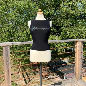 Svart ärmlös topp - Svart, tight och ärmlös topp från Koola Anna med färgglada små stenar i ett vågformat mönster över bröstet. Toppen har rund halsringning och är tillverkad i ett stretchigt, glansigt material som sitter snyggt mot kroppen.