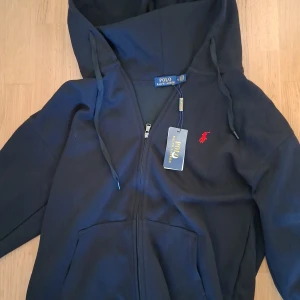 Svart hoodie från Polo Ralph Lauren XL - Svart hoodie från Polo Ralph Lauren i storlek XL. Klassisk design med dragkedja, huva med snörning och röd broderad logga på bröstet. Tillverkad i mjuk bomull och har praktiska fickor framtill. Perfekt för en chill och stilren look.