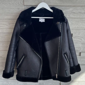 Svart pälsjacka från NA-KD, stl 34 - Snygg svart pälsjacka från NA-KD med stora slag, fuskpäls på krage och ärmslut samt dragkedjor på fickor och framtill. Fodrad med mjuk pälsimitation för extra värme.