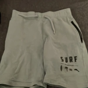 Ljusgröna shorts från Name It, strl 134 - Snygga ljusgröna shorts från Name It med tryck 'SURF SUMMER VIBES' och coola sommarikoner. Shortsen har snörning i midjan och en dragkedjeficka på sidan. Perfekta för chill dagar och strandhäng.