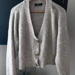 Grå kofta från Bershka med knappar - Mysig grå kofta från Bershka i stickat material. Koftan har v-ringning, tre stora knappar framtill och långa ärmar. Perfekt för lager-på-lager och chill dagar. Enkel att matcha med jeans eller kjol.