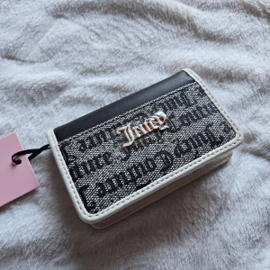 Juicy Couture plånbok i svart och beige - Juicy Couture plånbok i svart och beige med tryckt logga och textdetaljer. Utsidan har svart och grått tyg med mönster och metallogo, insidan har flera kortfack och dragkedjefack. Snygg och praktisk accessoar med coolt tryck.