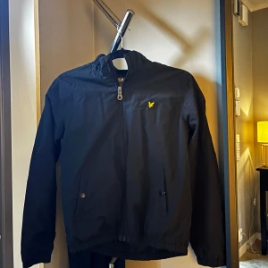 Svart vindjacka från Lyle & Scott - Svart vindjacka från Lyle & Scott med gul logga på bröstet. Jackan har dragkedja framtill, två fickor med knappstängning och resår vid ärmslut och midja. Perfekt för blåsiga dagar och enkel att matcha med det mesta.