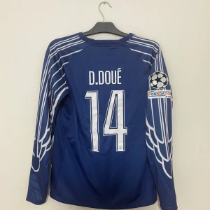 PSG långärmad blå fotbollströja D. Doué - Snygg blå PSG fotbollströja med vita detaljer och vingmönster på ärmarna. Långärmad modell med D. Doué och nummer 14 på ryggen, samt Champions League-märke och Qatar Airways-logga framtill. Tillverkad i lätt och ventilerande material.