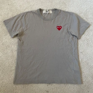 Grå t-shirt Comme des Garçons Play | S - Grå t-shirt från Comme des Garçons Play i storlek S. Klassisk rund hals och korta ärmar. På bröstet finns det ikoniska röda hjärtat med ögon, broderat. T-shirten är lite blekt på baksidan (syns mer på bild än i verkligheten)