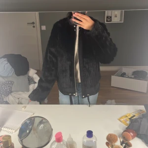 Svart pälsjacka med snörning - Säljer en svart pälsjacka med långa ärmar och cool snörning nertill. Jackan har en mjuk, fluffig känsla och stängs med dragkedja framtill. Perfekt för dig som vill sticka ut och hålla dig varm med stil.