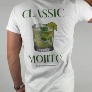 Classic Mojito - Låt sommaren leva vidare i varje steg med Classic Mojito – en avslappnad t-shirt inspirerad av friska jordgubbar, iskalla cocktails och lata dagar i solen.  Material: 100% mjuk och slitstark bomull Passform: Unisex, normal i storleken Färg: Klassisk vit med färgstarkt motiv Detalj: Tryck på ryggen, liten märkning nedtill