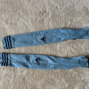 Ljusblå Adidas knästrumpor - Säljer ett par ljusblå knästrumpor från Adidas med mörkblå ränder upptill och Adidas-logga på framsidan. Strumporna har förstärkt häl och tå i mörkgrått samt mönstrad undersida. Perfekta till fotboll eller träning.