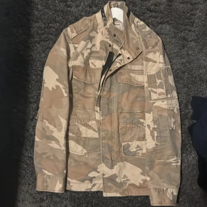 Camouflagejacka från Zadig & Voltaire - Snygg camouflagejacka från Zadig & Voltaire i beige och bruna toner. Jackan har flera fickor framtill, hög krage med tryckknappar och dragkedja. Tillverkad i bomull och har en cool militärinspirerad look. Perfekt för dig som vill sticka ut med streetstyle. Storlek M-L