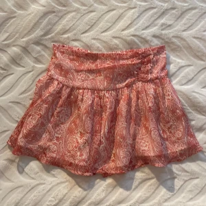 Rosa mönstrad kortkjol med volang - Supersöt kortkjol i rosa och vitt med paisleymönster och volangkant. Kjolen har bred resår i midjan och är gjord i ett lätt, luftigt material som passar perfekt till varma dagar. Mycket trendig. Priset kan diskuteras!💕