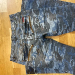 Blå camo jeans  - Säljer ett par blå jeans  med camouflagemönster och snygga guldfärgade dragkedjedetaljer på bakfickorna. bra skick fast gylfen är sönder