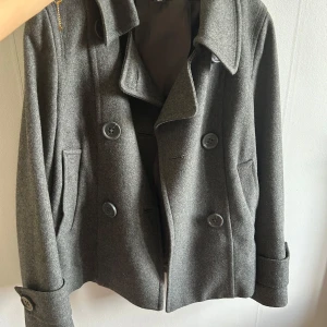 Grå kappa - Stilren grå skepparkavaj med dubbelknäppning och bred krage. Jackan har stora knappar framtill och dekorativa knappar bak, samt fickor på sidorna. Perfekt för dig som vill ha en klassisk och snygg look under höst och vinter.