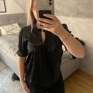 Svart blus i satin  - Säljer en svart blus i glansigt satinmaterial från JDY. Blusen har korta puffärmar, liten volangkrage och v-ringning framtill. Den är lös i passformen och har fina rynkade detaljer under bysten. Perfekt för dig som gillar en stilren och trendig look.