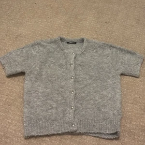 Grå stickad kofta från Gina Tricot - Mysig grå stickad kofta från Gina Tricot med korta ärmar och rund hals. Koftan har pärlknappar framtill och är tillverkad i ett fluffigt, mjukt material som känns extra skönt mot huden. Perfekt för lager-på-lager under kyliga dagar.