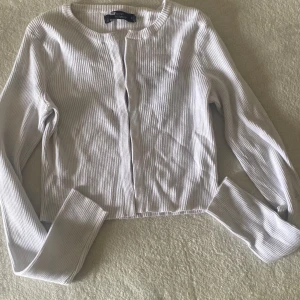 Vit ribbad kofta från New Yorker - Säljer en stilren vit ribbad kofta från Koton. Koftan har långärmad design, rund halsringning och är tillverkad i mjuk bomullsmix. Perfekt att slänga över en topp eller klänning för en enkel och fräsch look.