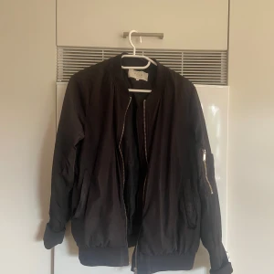 Svart bomberjacka från Vila, stl S - Svart bomberjacka från Vila med klassisk passform. Jackan har dragkedja framtill, ribbade muddar och fickor på sidorna samt en ärmficka med dragkedja. Perfekt för dig som gillar en stilren och enkel look.