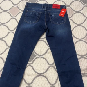 Mörkblå Dsquared2 jeans med slitningar - Snygga mörkblå jeans från Dsquared2 med coola slitningar och detaljer på både fram- och baksida. Klassisk femficksmodell med röd Dsquared2-lapp bak i midjan. Jeansen har raka ben och är tillverkade i mjukt denimtyg.