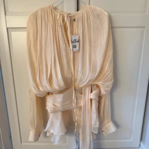 Champagnefärgad omlottklänning från Sella - Superfin kortklänning i champagnefärgad ton från Sella. Klänningen har omlott-detalj med knyt i midjan, volangkantad kjol och långa puffärmar med manschett och pärlknapp. Materialet är glansigt och skirt, vilket ger en lyxig känsla. Helt oanvänd + alla prislappar kvar!