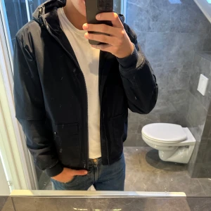 Mörkblå vindjacka med huva - Snygg mörkblå vindjacka med huva och dragkedja framtill. Jackan har två stora fickor med lock och knappar. Jag är 188cm och bär storlek M vilket passar bra. Pris kan alltid diskuteras! Hör av er vid frågor ;)