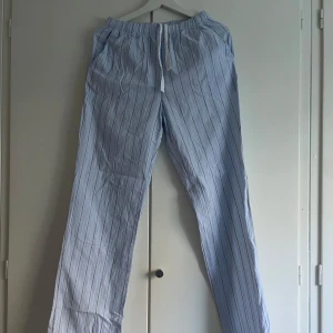 Ljusblå randiga linnebyxor med snörning - Snygga ljusblå linnebyxor med tunna vita ränder och bekväm snörning i midjan. Byxorna har en avslappnad passform och är perfekta för varma dagar. Materialet är lätt och luftigt, vilket gör dem sköna att bära.