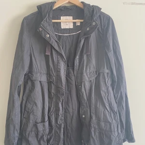 Svart parkajacka med huva - Svart parkajacka från H&M med huva och dragsko. Jackan har stora fickor framtill, dragkedja och knappar. Perfekt för kyliga dagar och har en avslappnad passform. Materialet är bomull och polyester.