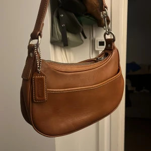 Vintage axelväska i skinn från Coach - Säljer en snygg brun axelväska från Coach i äkta skinn. Väskan har silverfärgade metalldetaljer, dragkedja upptill och en klassisk Coach-tag. Insidan har ett fack med dragkedja och väskan har en stilren, tidlös design som passar till allt. Denna väskan hittas ej i butik och har tyvärr inte kvitto den var köpt på marknad i barcelona.