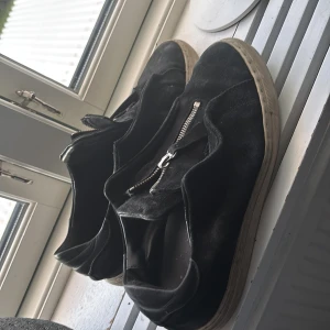 Axel arigato skor - Svarta sneakers i mocka med silvrig dragkedja framtill och vit sula. Skorna har en enkel och stilren design med rund tå och platt sula. Perfekta för dig som gillar en clean look med lite edge.
